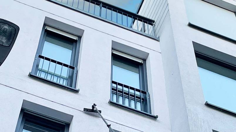 Ein modernes, mehrstöckiges Gebäude mit weißen Fassaden und großen Fenstern. Eine Teleskopstange mit Bürste wird zur Reinigung eines der oberen Fenster verwendet, während der Himmel blau und leicht bewölkt ist.