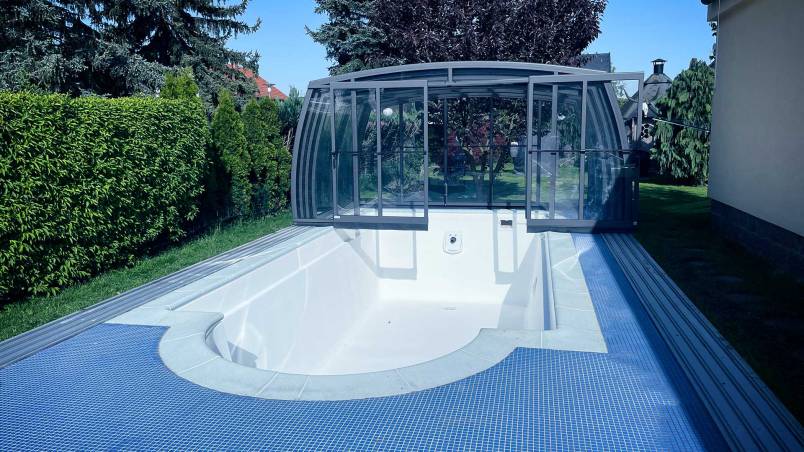 Ein leerer, weißer Swimmingpool mit blauer Fliesenumrandung und einer geschlossenen, modernen Poolüberdachung in einem gepflegten Garten mit grünen Bäumen und Sträuchern.